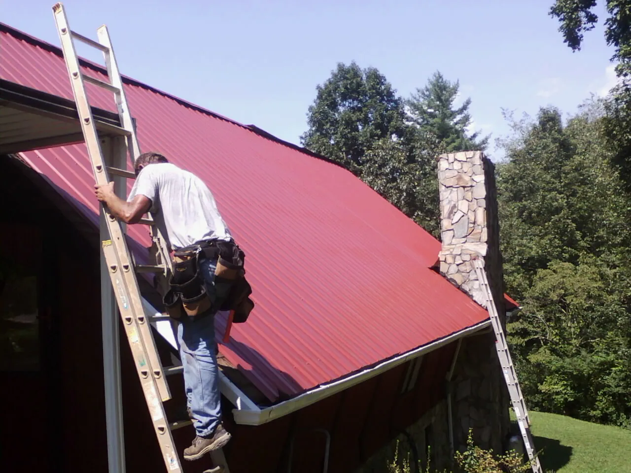 Roofer on a ladder beside a red metal roof and stone chimney — Soffit & Fascia Repair by Roofing Brothers in South Pomfret